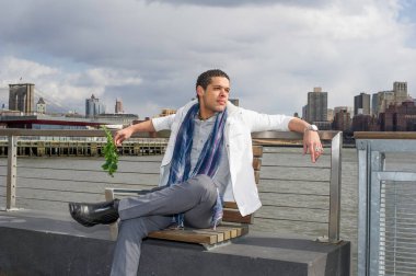 Büyük bir şehir hatlarına karşı, genç bir adam elinde yeşil yapraklarla nehrin kenarında oturuyor ve düşünüyor.