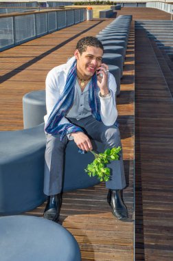 Elinde bir demet yeşil yaprak olan genç bir adam dışarıda modern bir bankta oturmuş telefonla konuşuyor.