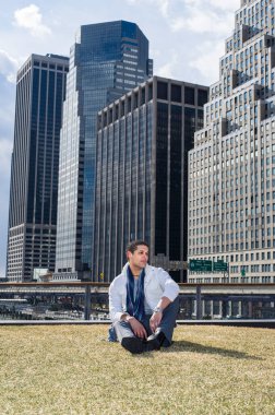 Genç bir öğrenci bir iş yerinin önündeki çimlerde oturuyor ve ileriye bakıyor.