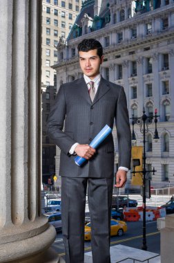 Genç bir işadamı bir ofis binasının önünde belgeleri tutuyor ve sana bakıyor.