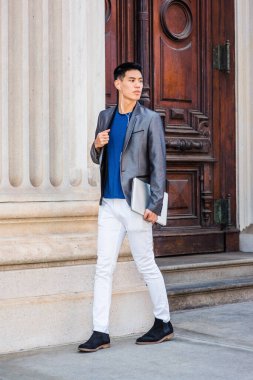 New York 'ta okuyan, gri ceket, beyaz pantolon, siyah ayakkabı giyen, kulaklık takan, dizüstü bilgisayar tutan, eski tarz ofis binasından çıkan genç Asyalı Amerikalı öğrenci.
