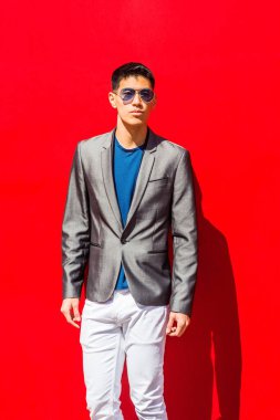 New York 'ta Asya kökenli genç bir adamın portresi, şık gri ceket, mavi atlet, beyaz pantolon, mavi güneş gözlüğü takıyor, güneşin altında kırmızı arka planda duruyor,
