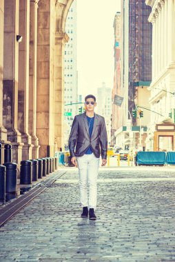 Genç Asyalı Amerikalı New York 'ta geziyor, gri ceket, beyaz pantolon, siyah ayakkabı giyiyor, güneş gözlüğü takıyor, kulaklık takıyor, müzik dinliyor, güneşli bir günde eski dar bir sokakta yürüyor.