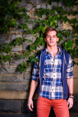 Mavi ve beyaz desenli gömlek, mavi kapüşonlu yelek, kırmızı kot pantolon giyen genç bir adam yeşil sarmaşık yapraklarıyla duvara dayanıyor, dinleniyor ve düşünüyor.