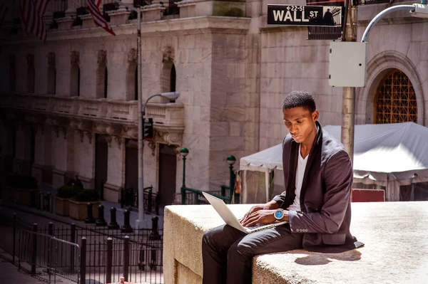 黒人の男が旅をしていて ニューヨークで働いていて ヴィンテージのオフィスビルのそばの通りに座っていて ノートパソコンで読み書きをしている 背景のウォール街の看板 — ストック写真