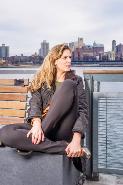 Güzel genç bir kadın nehir kenarında oturuyor, bacaklarını katlıyor ve ileriye bakıyor.