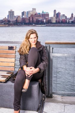 Güzel genç bir kadın nehir kenarında oturuyor, bacaklarını katlıyor ve sana gülümsüyor.