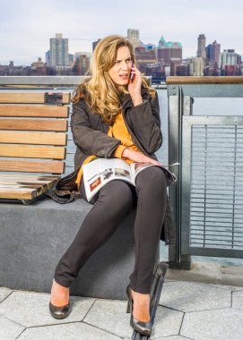 Kucağında bir dergi açarken, genç ve güzel bir kadın nehrin kenarında oturmuş, endişeli bir şekilde telefonla konuşuyor. Arka plan büyük bir şehir özeti..