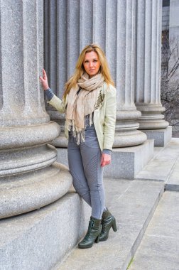Açık yeşil ceket, uzun bej eşarp, dar gri moda pantolon ve yeşil çizmeler giyen genç ve güzel bir kadın ofis binasının önünde bir sütunun yanında duruyor.