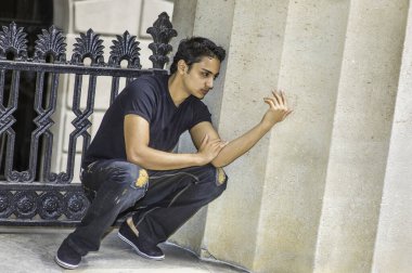 Siyah tişört ve modaya uygun bir pantolon giyen Asyalı genç, siyah metal korkuluk ve kolon arasında bir köşede çömeliyor ve derin düşüncelere dalıyor.