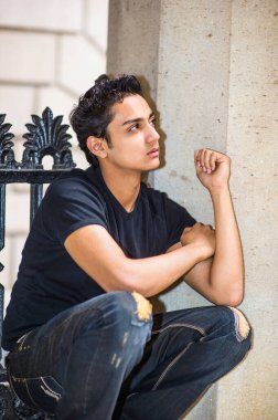 Genç Asyalı bir genç dışarıda çömeliyor, yukarı bakıyor ve düşünüyor..