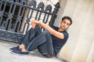 Çekici bir genç, siyah metal bir korkulukta oturuyor ve kendinden emin bir şekilde ileriye bakıyor.