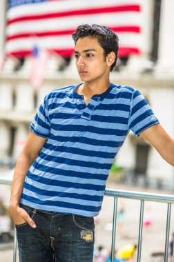 Şık bir pantolon ve elleri cebine koyan genç bir Asyalı genç, büyük bir sütunun yanında duruyor, aşağı bakıyor ve düşünüyor.