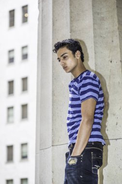 Şık bir pantolon ve elleri cebine koyan genç bir Asyalı genç, büyük bir sütunun yanında duruyor, aşağı bakıyor ve düşünüyor.