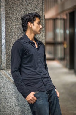 Siyah gömlek ve siyah pantolon giyen Asyalı genç, bir sütuna yaslanıyor ve derin düşüncelere dalıyor.