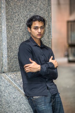 Siyah gömlek ve siyah pantolon giymiş, kollarını kavuşturmuş genç Asyalı bir delikanlı bir sütuna yaslanıyor ve derin düşüncelere dalıyor.