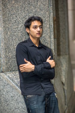 Siyah gömlek ve siyah pantolon giymiş, kollarını kavuşturmuş genç Asyalı bir delikanlı bir sütuna yaslanıyor ve derin düşüncelere dalıyor.