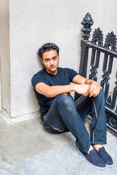 Siyah tişört ve şık bir pantolon giyen Asyalı genç, siyah metal bir korkulukta oturuyor ve sana bakıyor.