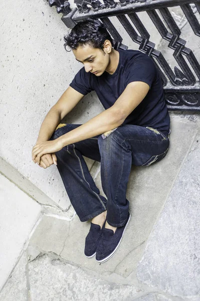 Siyah tişört ve şık bir pantolon giyen Asyalı genç, siyah metal bir korkuluğun yanında oturuyor ve derin düşüncelere dalıyor.