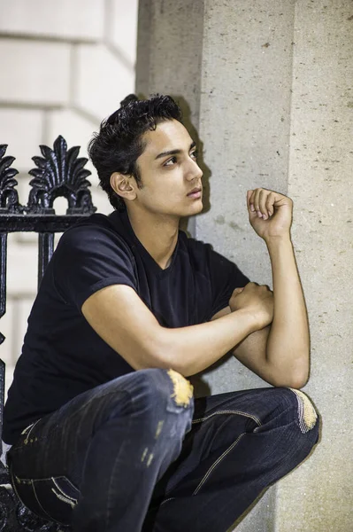 Genç Asyalı bir genç dışarıda çömeliyor, yukarı bakıyor ve düşünüyor..