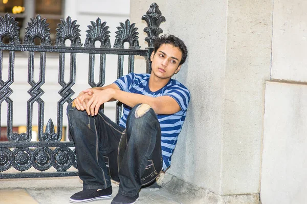 Mavi çizgili bir tişört ve şık bir pantolon giyen Asyalı genç, siyah metal bir korkuluk ve duvar arasında, derin düşüncelere dalmış bir şekilde çömeliyor.