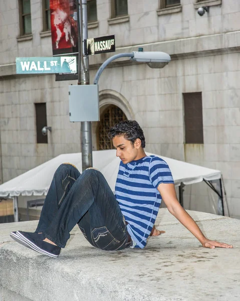 Mavi çizgili bir tişört, şık bir pantolon ve caddenin kenarındaki bir sahnede oturan Asyalı bir genç, aşağıya bakıyor ve düşünüyor.