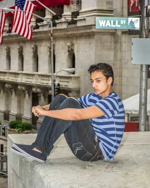 Mavi çizgili bir tişört, şık bir pantolon ve caddenin kenarındaki bir sahnede oturan Asyalı bir genç, aşağıya bakıyor ve düşünüyor.