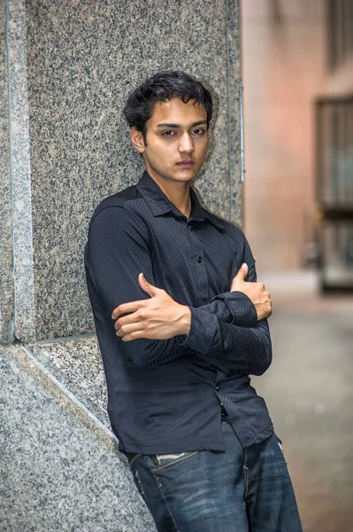 Siyah gömlek ve siyah pantolon giymiş, kollarını kavuşturmuş genç Asyalı bir delikanlı bir sütuna yaslanıyor ve derin düşüncelere dalıyor.