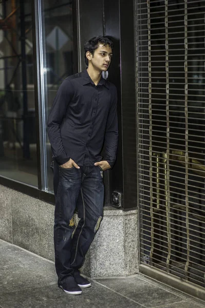 Siyah gömlek ve şık bir pantolon giyen genç Asyalı bir genç, kapalı bir kapıya yaslanıyor, içine bakıyor ve düşünüyor.