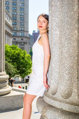 Beyaz kolsuz iş kıyafetleri giyen profesyonel bir kadın, ofis binasının dışındaki bir sütuna yaslanıyor, rahatlıyor ve düşünüyor.