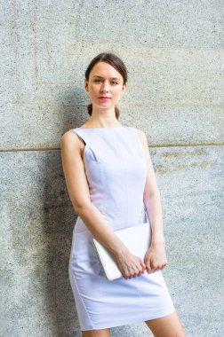 Beyaz kolsuz iş kıyafetleri giymiş, elinde küçük bir bilgisayar olan bir iş kadını, dışarıda durmuş, düşünceli bir şekilde sana bakıyor.