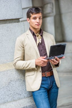 Bej renkli bir ceket, kahve rengi bir gömlek, mavi kot pantolon, boynuna bağlanmış bir eşarp ve elinde küçük bir bilgisayar olan yakışıklı genç bir adam düşünceli bir şekilde ileriye bakıyor..