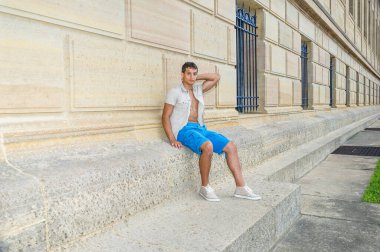 Demir kısa kollu gömlek, mavi şort ve spor ayakkabı giyen genç ve çekici bir adam bir desen duvarına yaslanıp dinleniyor..