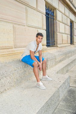 Demir kısa kollu gömlek, mavi şort ve spor ayakkabı giyen genç ve çekici bir adam, bir şablon duvarın yanında oturuyor, rahatlıyor ve düşünüyor..