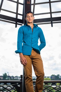 Mavi gömlekli, kahverengi pantolonlu, genç ve çekici bir adam gökyüzünün önünde duruyor ve içtenlikle sana bakıyor..