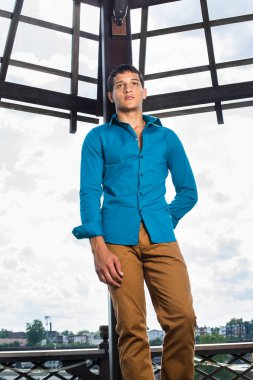 Mavi gömlekli, kahverengi pantolonlu, genç ve çekici bir adam geniş gökyüzünün önünde duruyor ve sadakatle ileriye bakıyor..