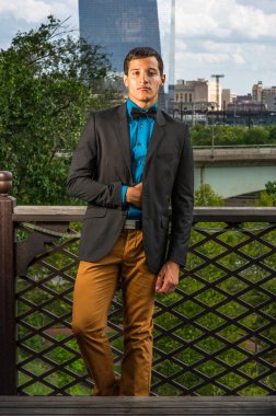 Siyah bir ceket, mavi bir gömlek ve siyah bir papyon giyen genç bir işadamı dışarıda duruyor ve kendinden emin bir şekilde ileriye bakıyor. Arka plan modern bir iş binasıdır.