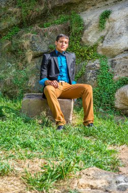 Siyah ceket, mavi gömlek, kahverengi pantolon ve siyah papyon giyen genç bir iş adamı, kayaların üzerinde oturup düşünüyor.