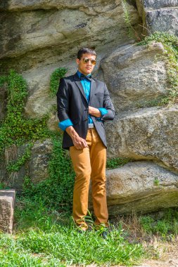 Siyah ceket, mavi gömlek, kahverengi pantolon ve siyah papyon giyen, güneş gözlüğü takan genç bir işadamı dışarıda durup mola veriyor..