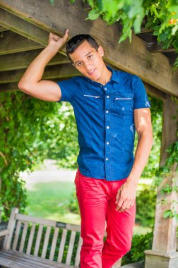 Mavi kısa kollu gömleği ve kırmızı pantolonu olan yakışıklı, çekici bir adam ahşap bir yapının yanında duruyor ve yeşil yapraklar, büyüleyici bir şekilde sana bakıyor.