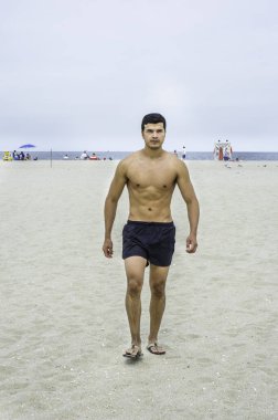 Genç, erkeksi bir adam, yarı çıplak, sahilde yürüyor..