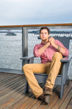 Açık kırmızı gömlekli, koyu sarı pantolonlu ve kahverengi ayakkabılı genç yakışıklı bir adam nehir kıyısındaki bir sandalyede oturuyor, ellerini sıkıca tutuyor, ayak basıyor, dinleniyor..