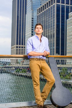 Açık mavi gömlekli ve koyu sarı kotlu genç, enstrüman kutusu olan yakışıklı bir müzisyen iş yerinin önünde kendinden emin bir şekilde bekliyor..