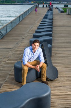 Açık mavi gömlek, koyu sarı kot pantolon ve kahverengi ayakkabı giyen, müzik aleti kutusu olan yakışıklı genç bir müzisyen akşamları modern bir bankta oturuyor, büyüleyici bir şekilde sana bakıyor..