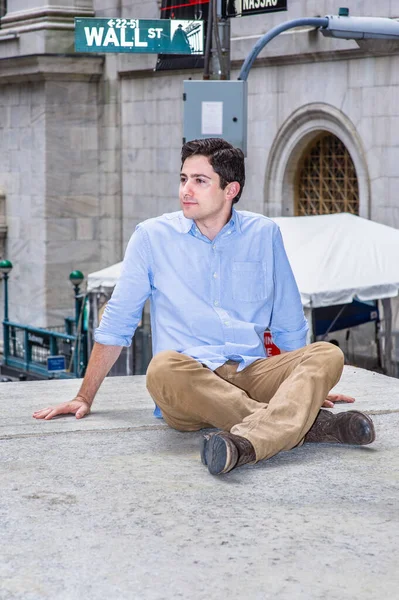 Genç yakışıklı bir adam caddenin köşesinde bir sahnede oturuyor, rahatlıyor ve düşünüyor. Arka planda Wall Street tabelası var..