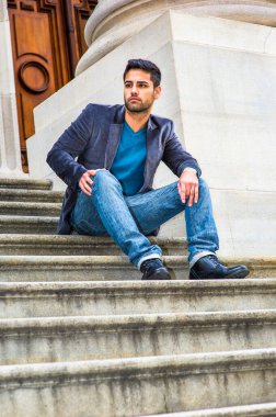 Koyu mor yün ceket, mavi kot pantolon ve siyah deri ayakkabılar giyen, ekmek ve bıyıklı genç bir Orta Doğulu kapı eşiğinde oturmuş, rahatlıyor ve düşünüyor..