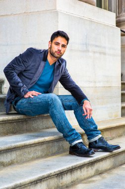 Koyu mor yün ceket, mavi kot pantolon ve siyah deri ayakkabılar giyen, ekmek ve bıyıklı genç bir Orta Doğulu, düşünceli bir şekilde sana bakıyor.