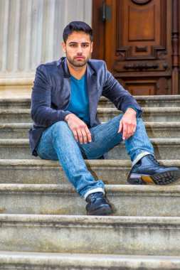 Koyu mor yün ceket, mavi kot pantolon ve siyah deri ayakkabılar giyen, ekmek ve bıyıklı genç bir Orta Doğulu kapı eşiğinde oturuyor, yorgun.