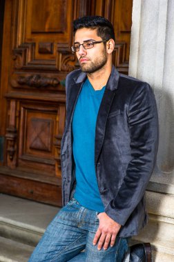 Koyu mor yün ceket, kazak ve kot pantolon giyen, gözlük takan, ekmek ve bıyıklı genç bir alim, nedense derin düşüncelere dalmak için kapının önünde duruyor.