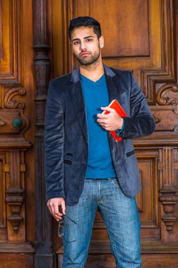 Yünlü bir ceket giymiş, kazak ve kot pantolon giymiş, elinde gözlük ve kırmızı bir kitap tutan, ekmek ve bıyıklı genç ve yakışıklı bir profesör, ofisin kapısında duruyor, düşünceli bir şekilde sana bakıyor.